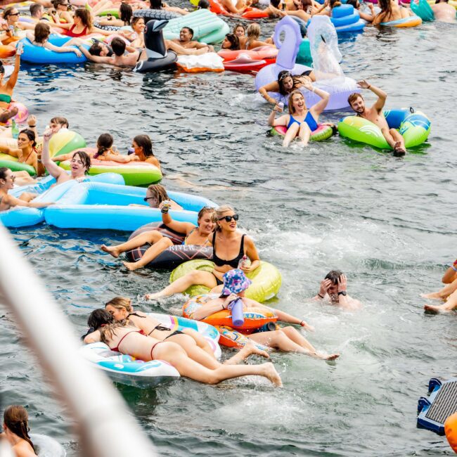 People enjoying a sunny day in the water with colorful inflatable floats, including unicorns and donuts. They're near a boat, and everyone appears relaxed and cheerful. The atmosphere is lively and festive.