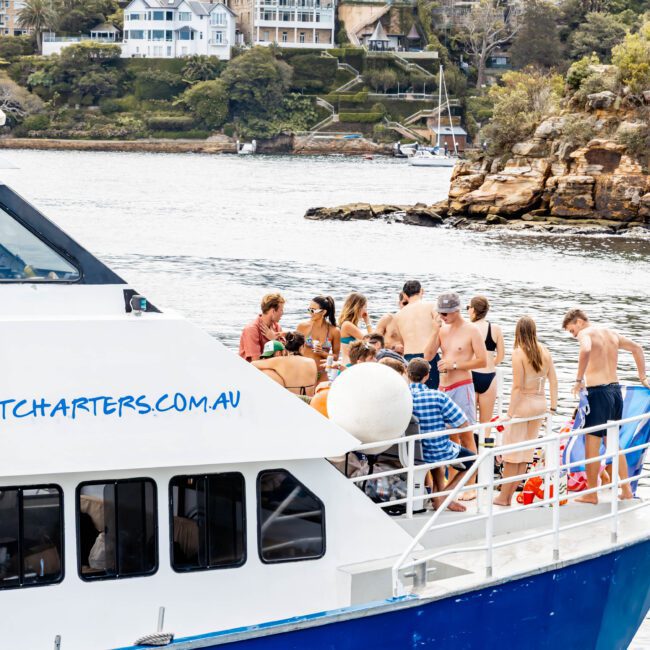 A group of people in swimsuits and casual attire socializing on the deck of a blue and white yacht. The yacht is sailing near a rocky shoreline with trees and buildings in the background. The water is calm under a cloudy sky.