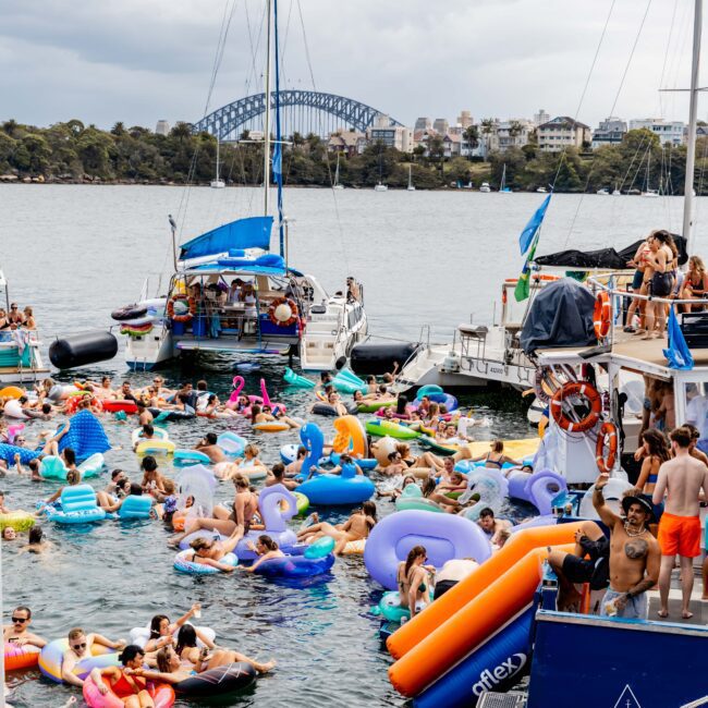 A lively gathering on the water features several people on colorful inflatables and boats. In the background, a large bridge spans the skyline with buildings nestled in greenery. Cloudy skies loom overhead.