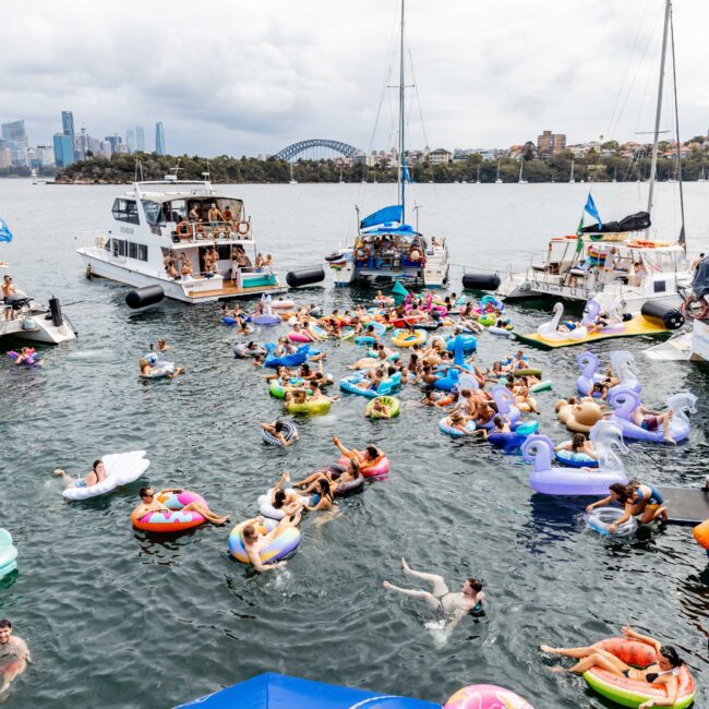 A group of people relax on colorful inflatables in a harbor surrounded by boats. The sky is cloudy, and a city skyline is visible in the background. Some boats display flags, and a few attendees are on board, enjoying the event.