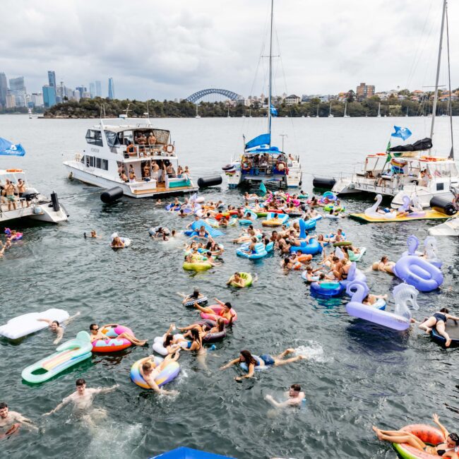 A large group of people enjoys a pool party in a body of water, using inflatable floats. Boats surround them, and a city skyline and bridge are in the background. The scene is festive and crowded.