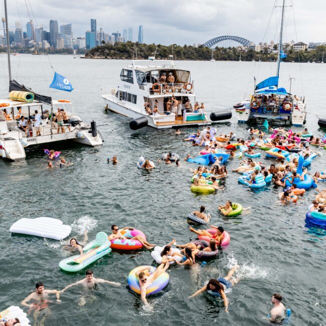 A lively scene of people on colorful inflatables in the water, near several boats. The background shows a city skyline and a large bridge. The atmosphere is festive and crowded, with a partly cloudy sky overhead.