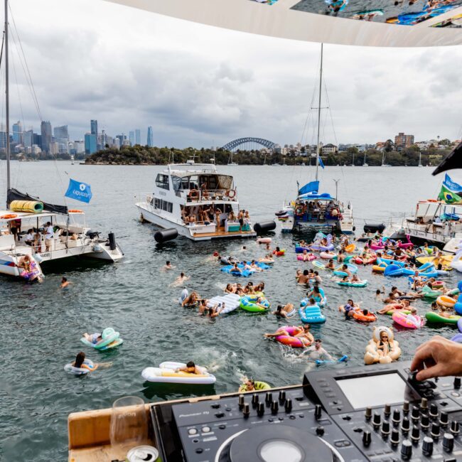A DJ performs on a yacht overlooking a lively party on the water. People float on colorful inflatables near several boats. The skyline and bridge are visible in the cloudy background.