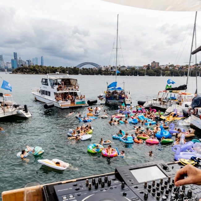 View from a DJ booth on a yacht, overlooking a lively party on a harbor. Many people are on colorful inflatables in the water. Boats and the city skyline, including a bridge, are visible in the background under a cloudy sky.
