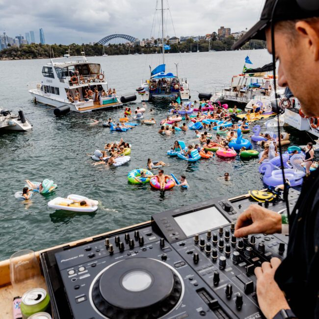A DJ mixes music on a yacht overlooking a vibrant pool party with people on colorful inflatables in the water. Boats surround the gathering, and a cityscape with a bridge is visible in the background under a cloudy sky.