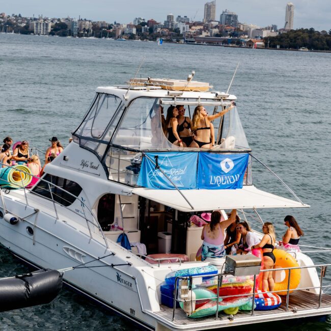 A group of people in swimwear are enjoying a party on a white yacht in a city harbor. The yacht is decorated with colorful pool floats and a blue banner. In the background, city buildings and a clear sky are visible.