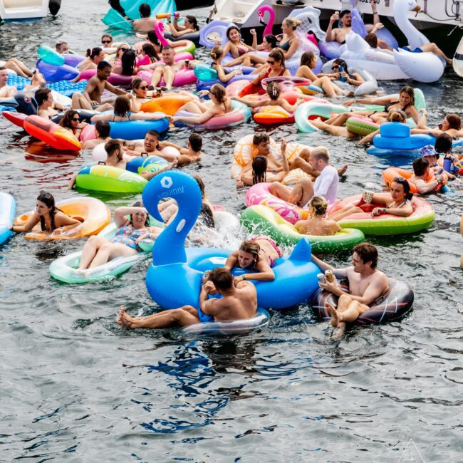 A lively group of people enjoying a sunny day on a lake, floating on various colorful inflatable rafts and pool toys. Some are on flamingos, unicorns, and other fun shapes, near several docked boats. Everyone seems to be relaxed and having a good time.