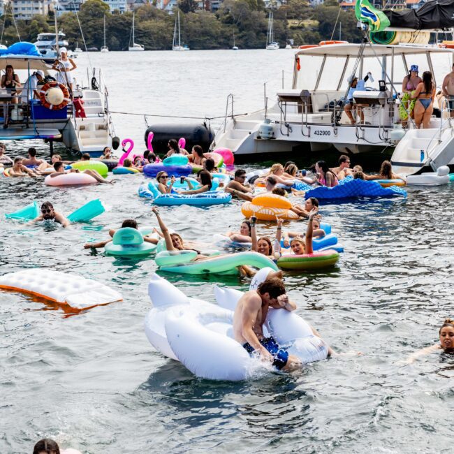 People enjoying a sunny day on a river with colorful inflatables, including a white swan. Two boats are docked nearby, and people are swimming and lounging. Trees and buildings are visible in the background.