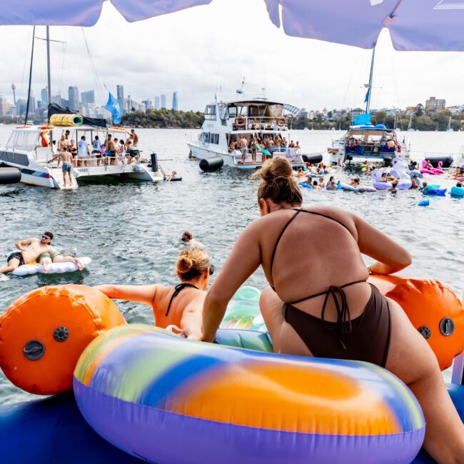 People enjoy a sunny day on the water, swimming and lounging on colorful inflatables near several boats. The city skyline is visible in the background, and a person holds a water bottle on a boat's deck.