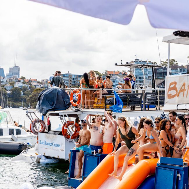 People enjoying a sunny day on a boat with water slides. Some are using the slide, while others relax on the deck. The boat is anchored, and there are inflatable floats in the water. Buildings and trees are visible in the background.
