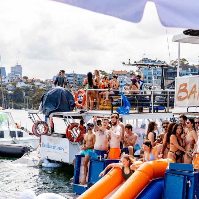 A group of people enjoying a sunny day on a boat party. Some are lounging on inflatables in the water, while others socialize on the deck. The backdrop shows a cityscape and more boats, hinting at a festive atmosphere.