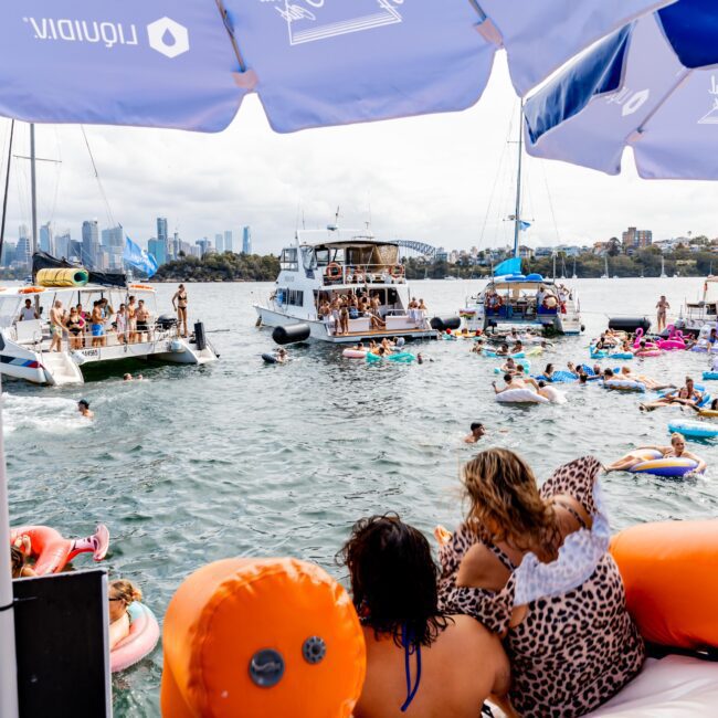 People enjoy a social event on a harbor, surrounded by boats and floating inflatables. In the background, a city skyline is visible under a cloudy sky. The scene is lively and festive, with groups gathered on vessels and in the water.