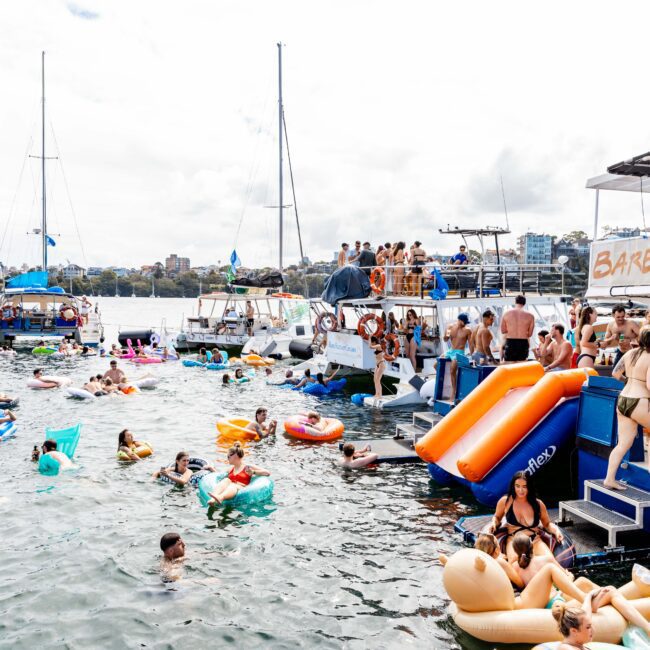 A lively scene of people enjoying a sunny day on the water with several boats, inflatables, and swimmers. A large boat with a slide labeled "Barefoot Blue" is to the right, and a bridge is visible in the background.
