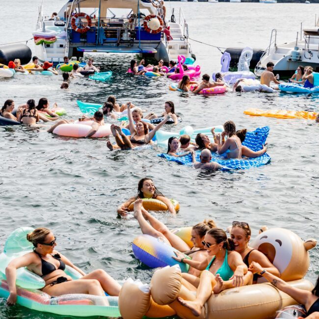 People relax on inflatable floats in a busy waterway, surrounded by boats. They appear to be enjoying a sunny day. A bridge and city buildings are visible in the background.