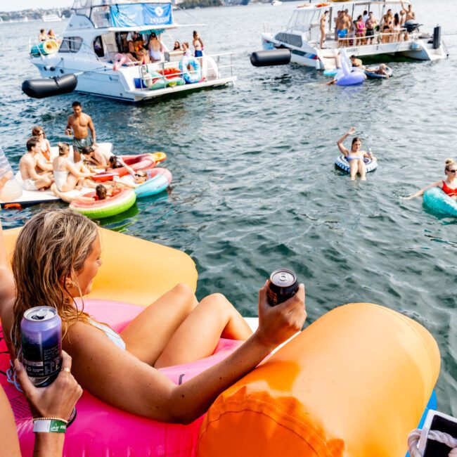 People enjoying a sunny day on the water, lounging on colorful inflatable floats with drinks in hand. Boats are anchored nearby, and the city skyline is visible in the background. A lively, festive atmosphere.