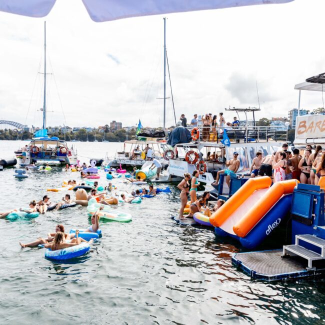 A lively water party featuring people on inflatable floats and pool toys in a sunny bay. Several boats are docked, including one with an inflatable slide. Groups of people are swimming and socializing in the water. A bridge and buildings are visible in the background.