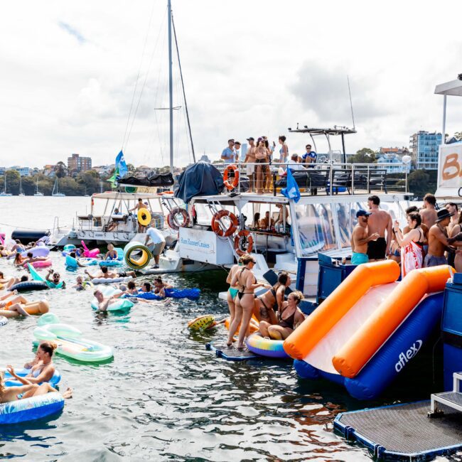 A lively scene with people enjoying a party on anchored boats and inflatables in the water. The area is bustling with activity as attendees socialize, swim, and relax on colorful floaties under a partly cloudy sky.