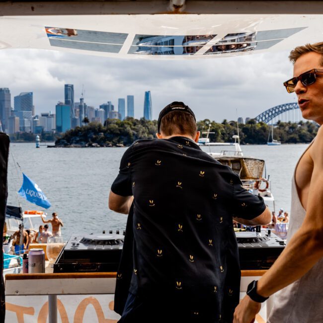 Two people on a boat, one DJing with equipment, wearing a black shirt and cap. The other in sunglasses and a sleeveless shirt looks away. Behind them, a city skyline, water, and Sydney Harbour Bridge are visible.