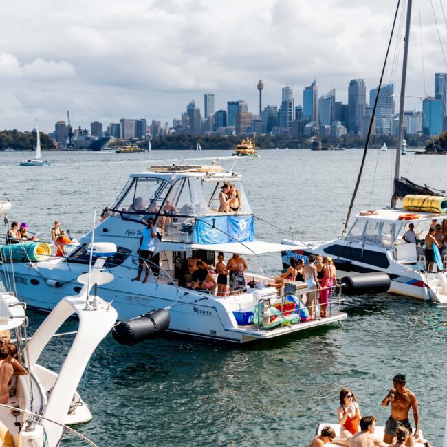 Boats anchored in a harbor with people sunbathing and socializing aboard. The city skyline is visible in the background under a cloudy sky.