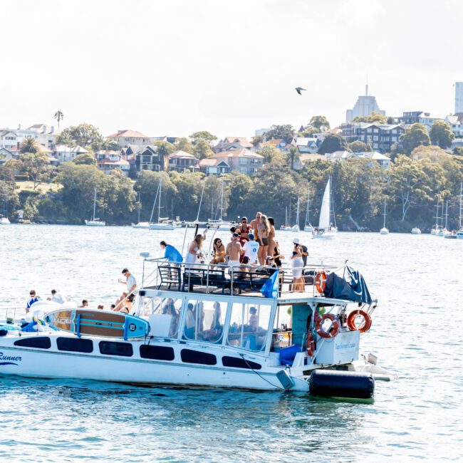 A group of people are gathered on the deck of a small boat floating on a sunny body of water. Sailboats are visible in the background, with a distant view of houses on a tree-lined shore.