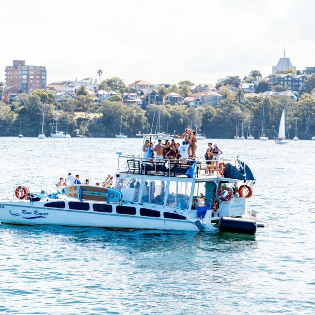 A group of people are enjoying themselves on a boat named Ren Runner in a sunny harbor. The boat is surrounded by calm water, with sailboats and a cityscape in the background. The sky is clear.