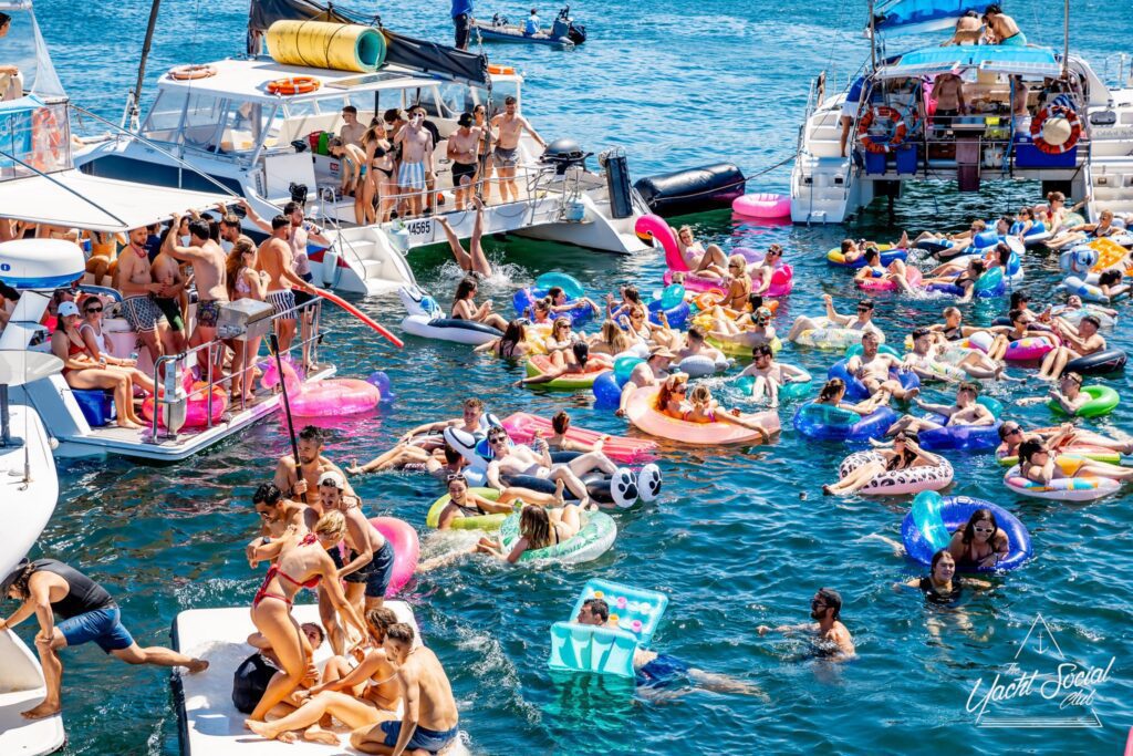 A lively scene of people partying on yachts and colorful inflatables in the water, enjoying a sunny day. The crowd is having fun, swimming, lounging, and socializing in a festive atmosphere.