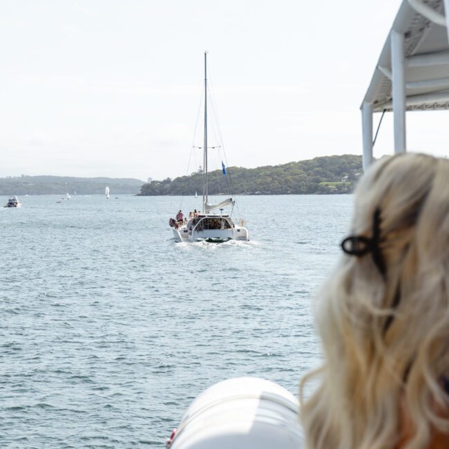 A sailboat glides on the water with two people visible on deck. In the foreground, a person with wavy blond hair and sunglasses looks towards the boat. A distant shoreline is visible under a cloudy sky.