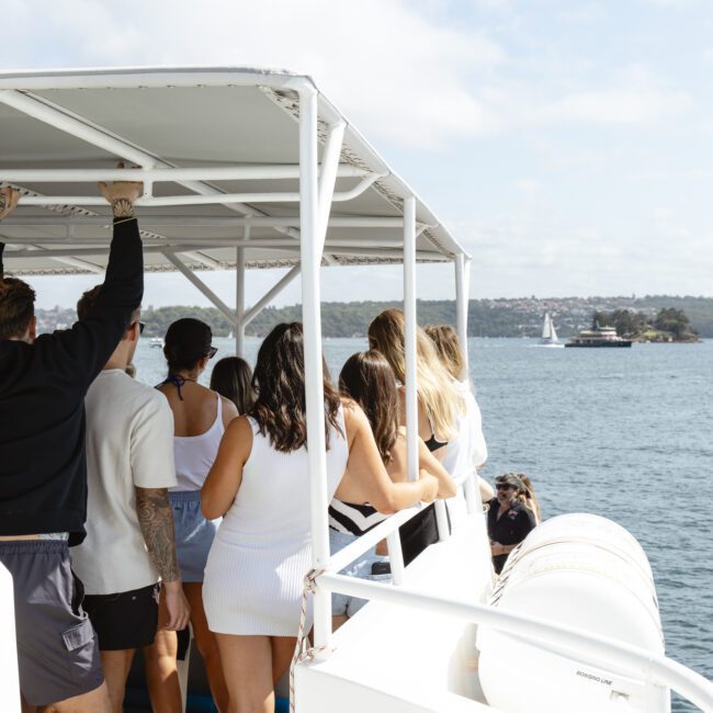 A group of people stand on a boat deck, enjoying a sunny day. The boat sails on calm water with a distant shoreline and a few sailboats visible. The sky is partly cloudy, creating a relaxed atmosphere.