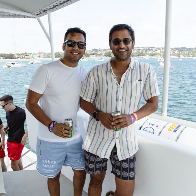Two men are smiling and wearing sunglasses while standing on a boat. One is in a white t-shirt and blue shorts, and the other is in a striped shirt and shorts. They are holding drinks, with a scenic ocean view and other boats in the background.