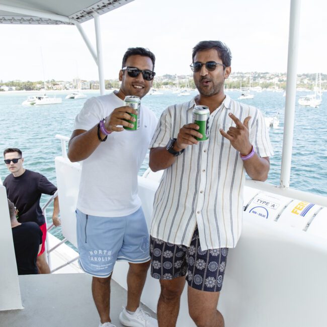 Two people are standing on a boat, holding drinks and smiling. One wears a white T-shirt and blue shorts, the other a striped shirt and patterned shorts. They are by a railing with water and boats in the background.