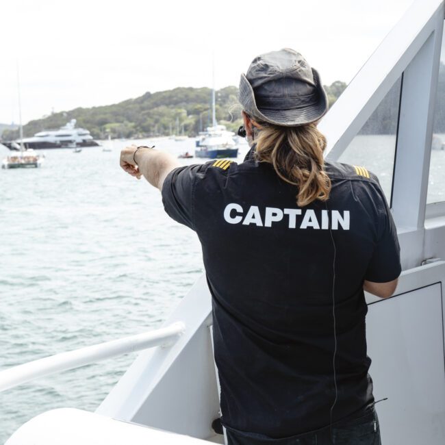 A person wearing a black shirt with "CAPTAIN" printed on the back stands on a boat, pointing towards the water. Several yachts are visible across the water, with a shoreline and trees in the background.