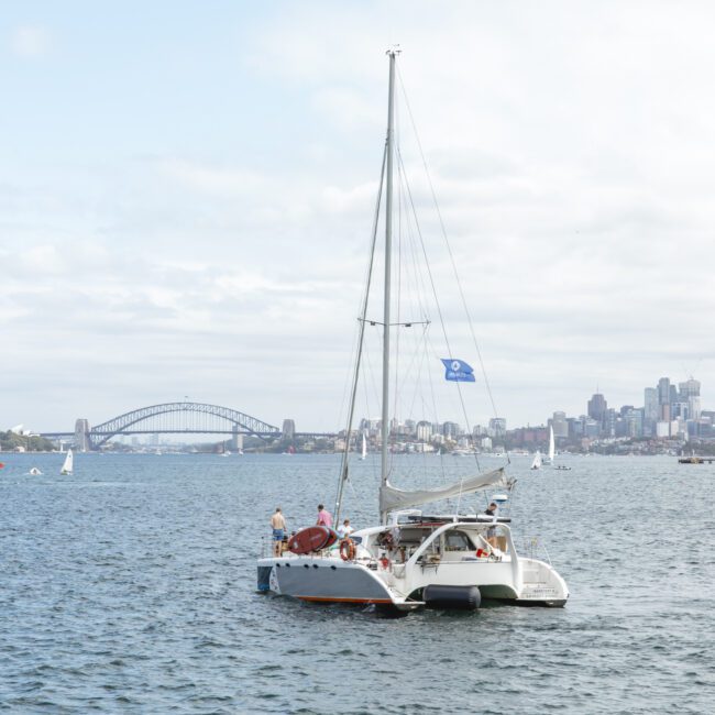 A catamaran sails on a calm body of water. The backdrop includes a city skyline with skyscrapers and a prominent bridge under a partly cloudy sky. Several smaller boats are visible in the distance.