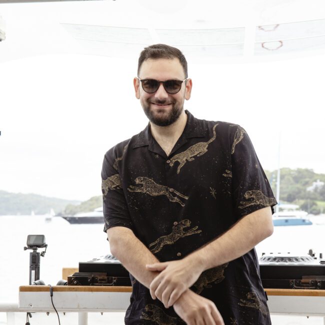 A man with a beard, wearing sunglasses and a black shirt with a crocodile pattern, stands on a boat with DJ equipment. The background features a scenic view of water and distant sailboats on a cloudy day.
