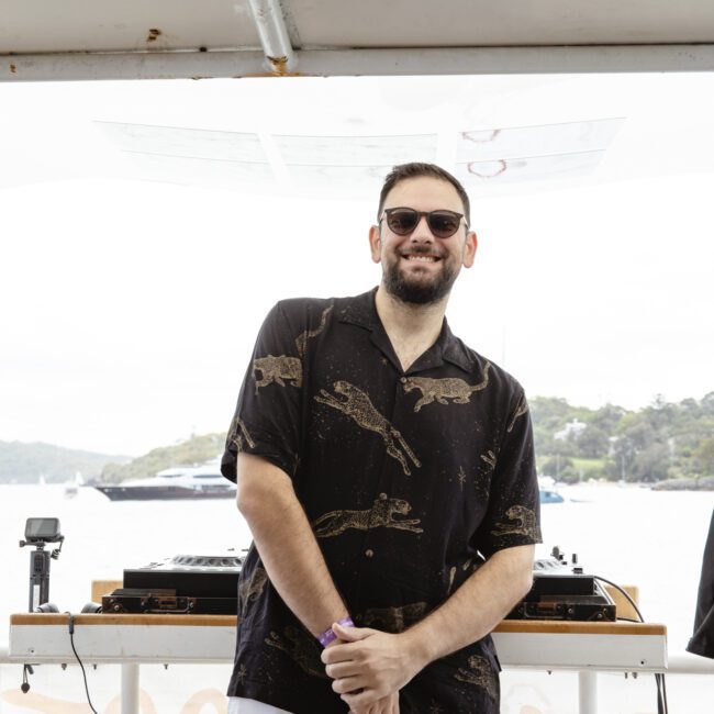 A person wearing sunglasses, a dark patterned shirt, and white pants stands smiling on a boat. DJ equipment is visible behind them. The background shows a body of water and a partially cloudy sky.