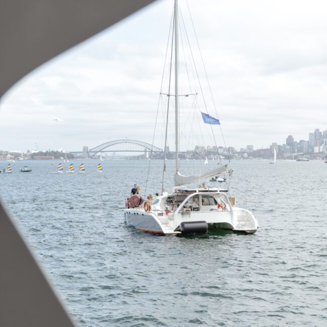 A white sailboat with passengers is on a body of water. The Sydney Harbour Bridge is visible in the background. The photo is framed by a triangular shape in the foreground. The sky is cloudy.