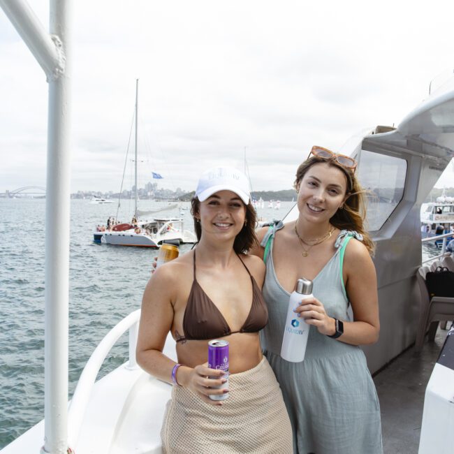 Two women smiling on a boat, each holding a beverage. They are standing near the railing with a view of water and several boats in the background. One wears a brown bikini top and a cap, the other a light blue dress.