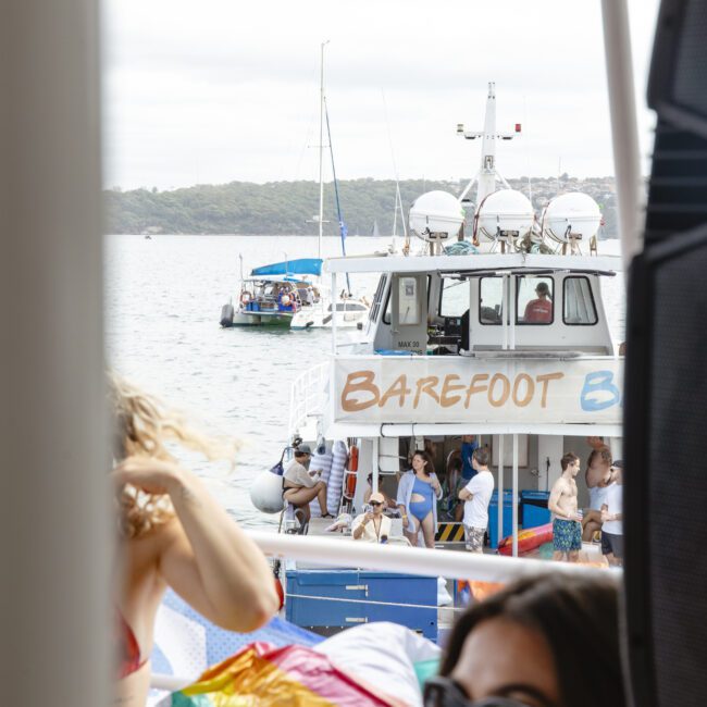 People are relaxing on a boat with a rainbow flag visible. Another boat labeled "Barefoot" is in the background on the water, with multiple people on board. The sky is overcast.