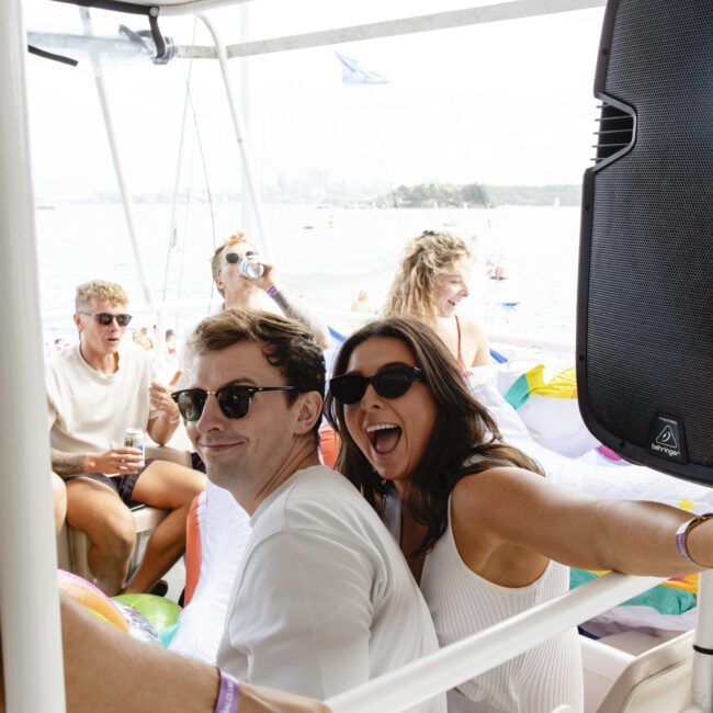 A group of people enjoying a sunny day on a boat. They are casually dressed in summer outfits, some wearing sunglasses. The scene is lively, with smiles and laughter, and a clear view of the water and distant shoreline in the background.