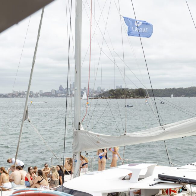 A group of people in swimwear are on a yacht with a life preserver on deck. A blue flag waves above. The background shows a city skyline under a cloudy sky, and other small boats are visible on the water.