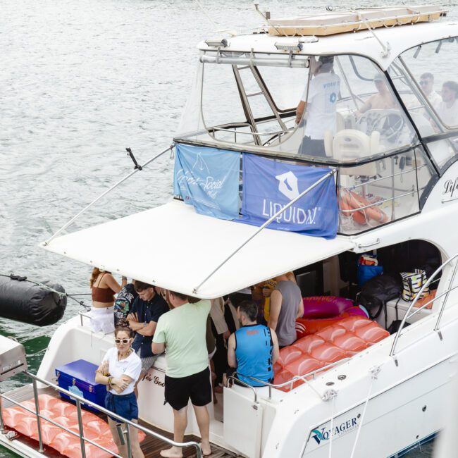 A group of people on a yacht with a "Liquid" banner, docked near a forested shoreline. They are socializing on the deck with orange life jackets visible. The setting suggests a relaxed gathering on the water.