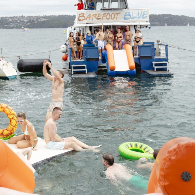 A group of people enjoying a day on the water. Some are on a boat named "Barefoot Blue," featuring orange slides. Others are swimming and lounging on inflatables, including a green inner tube. The sky is cloudy, and land is visible in the background.