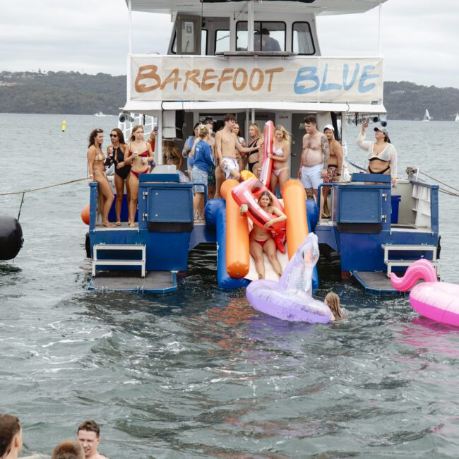 A group of people in swimsuits are having fun on the back of a boat named "Barefoot Blue." Some hold inflatable pool toys, and others enjoy the water. The boat is on a body of water with a shoreline visible in the background.