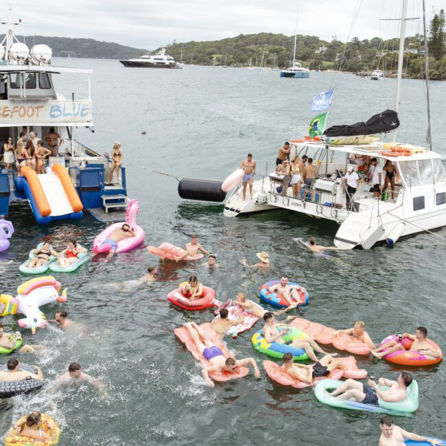 A lively scene on the water with two anchored boats. People are floating on colorful inflatables, like unicorns and pizzas. The boat named "Barefoot Blue" is visible, and the setting is surrounded by trees and distant yachts.