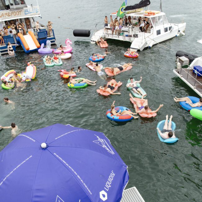 A group of people are floating on colorful inflatable rafts in a body of water near two boats. One boat has a slide, and an umbrella with "Yacht Social Club" branding is visible in the foreground. There are trees and hills in the background.