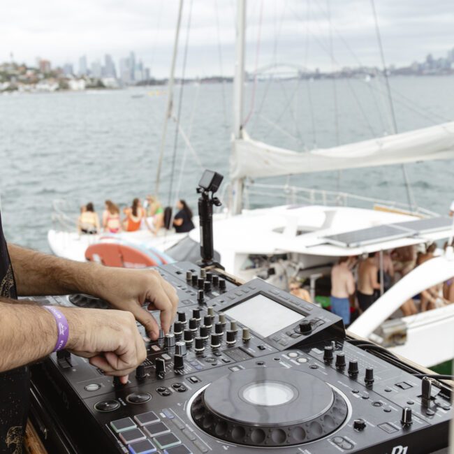 A DJ adjusts controls on a Pioneer DJ console aboard a yacht. The background shows the ocean and blurred people on nearby boats with a city skyline in the distance.