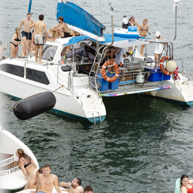 A group of people relax and socialize on a sailboat and a floating platform in the water. Some are sunbathing, while others chat on the boat. The backdrop features a calm body of water and distant hills.