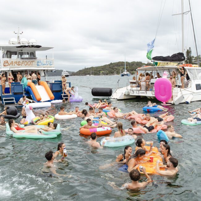 A lively gathering of people enjoying a day on the water, floating on colorful inflatables between two boats. The sky is overcast, and the mood is festive with drinks being shared. The backdrop features a hilly landscape.