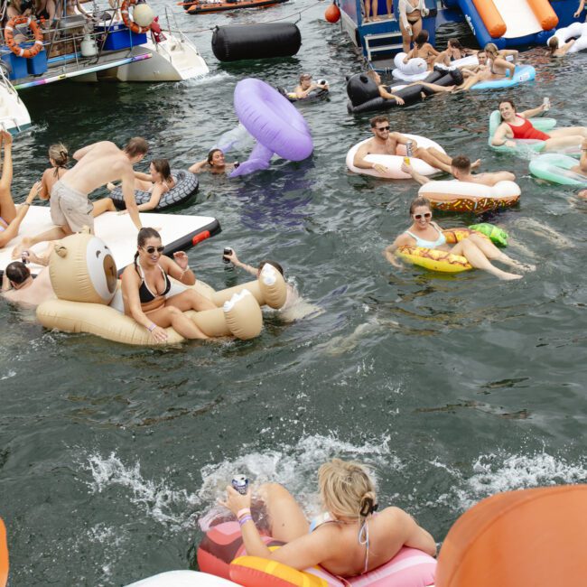 A lively scene with people relaxing on colorful inflatables in the water near boats. The group is enjoying a sunny day, lounging on various pool floats, including a bear-shaped float and a pretzel-shaped one, with a backdrop of a partially cloudy sky.