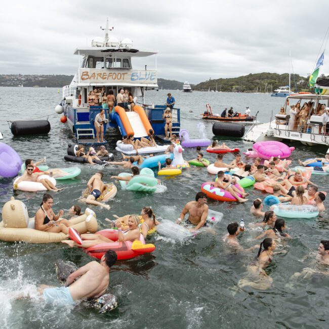 A large group of people floats on colorful inflatables in the water, near boats. A boat with "Barefoot Blue" has slides leading to the water. Other boats are nearby. It's a lively scene with people enjoying a day on the water under a cloudy sky.