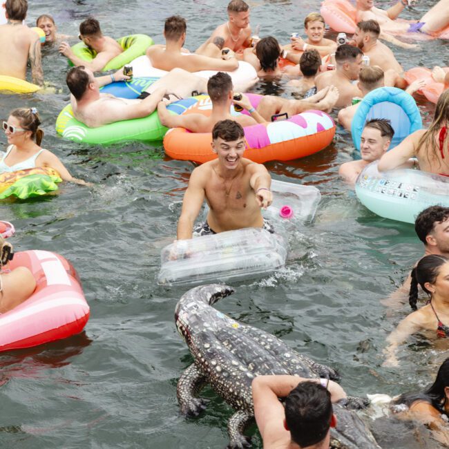 People enjoying a water party, floating on colorful inflatable tubes and rafts, including a unicorn and alligator. One person is holding a cooler. It's a sunny, lively scene with many having fun in the water.